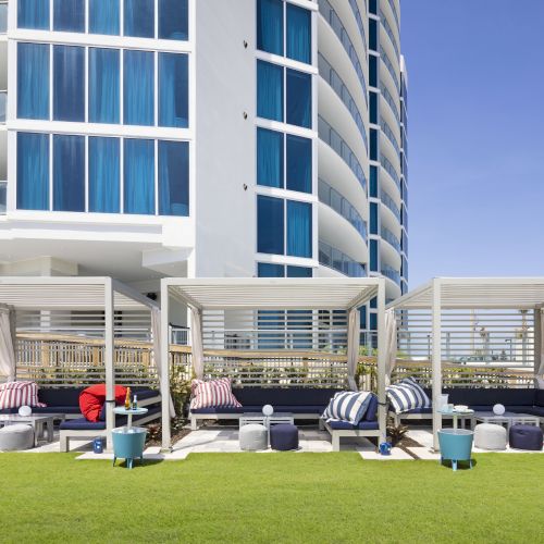 The image shows a modern outdoor lounge area with cabanas, furniture, and grass in front of a multi-story building with blue windows, under a clear sky.