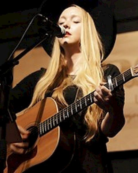 A person with long blonde hair is playing an acoustic guitar and singing into a microphone on stage, framed by a dark background.
