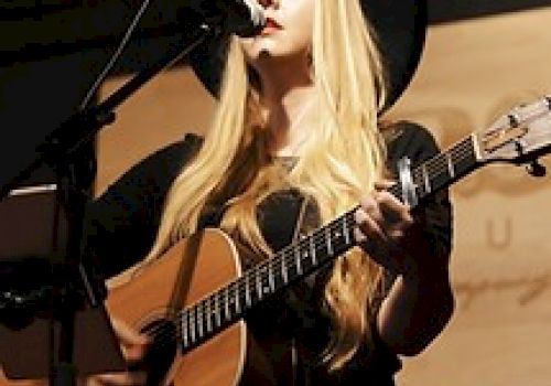 A person with long blonde hair is playing an acoustic guitar and singing into a microphone on stage, framed by a dark background.