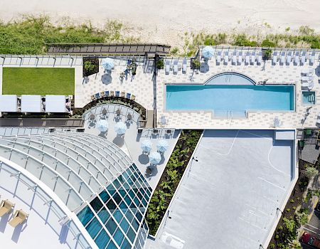 An aerial view of a modern resort featuring a swimming pool, sun loungers, a green lawn area, and a nearby beach, with part of a glass dome visible.