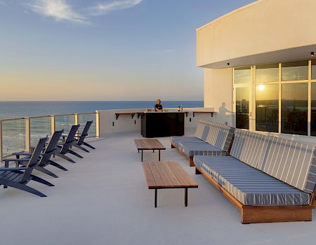 A rooftop terrace with ocean views, featuring lounge chairs, striped cushioned seating, tables, and a bar area with a person standing behind it.