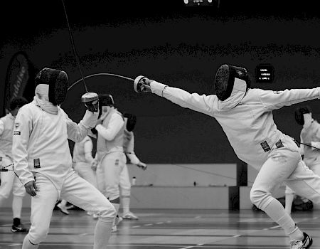 Two fencers in action during a match, wearing protective gear and masks, swords crossed, with others visible behind them.